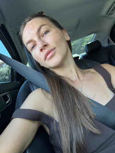A woman is sitting in the passenger seat of a car, likely in Prijeradi, Montenegro. She is wearing a brown dress and a seatbelt. Her hair is long and brown, and she is wearing a necklace with a diamond pendant. The background shows trees and greenery, suggesting the car is moving through a suburban or rural area. The lighting indicates it is daytime.