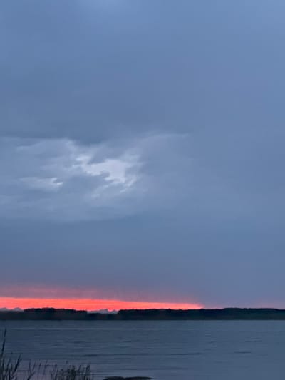 The image depicts a wide, calm body of water, likely a lake, reflecting the sky. A thin strip of land, possibly a shoreline with vegetation, divides the water from the sky. The sky is a muted blue, transitioning to a vibrant, fiery orange-red glow along the horizon, suggesting either a sunrise or sunset. Wispy clouds are visible in the sky, illuminated by the warm light. The overall mood is serene and tranquil, with a sense of natural beauty. There is no visible text in the image. The location is Aiyrtau, Kazakhstan.