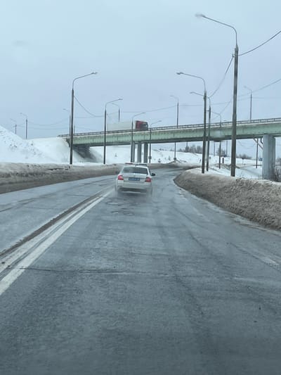 The image depicts a winter or early spring road scene in the city of Ivashevo, Russia, under an overcast sky. A two-lane asphalt road, partially wet with visible puddles and worn double solid white lines, recedes into the distance. A white sedan, identifiable as a police or emergency vehicle by its blue and white checkered roof markings and red light bar, travels away from the viewer in the right lane, leaving a slight water spray from its rear wheels. Its Russian license plate, "P 309 XA 90," is partially visible. Snowbanks, showing signs of melting and dirt, line both sides of the road. Multiple utility poles with streetlights and overhead wires extend along the right side of the road. In the background, an elevated overpass, supported by concrete pillars, crosses above the road. A red semi-truck with a white trailer is observed driving on this overpass. Beyond the overpass, additional snow-covered terrain and sparse, bare trees are visible.