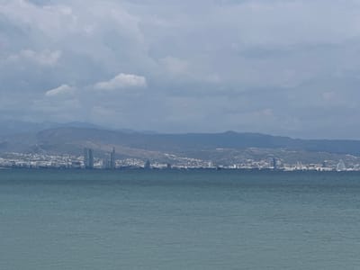A distant coastal city, identified as the city of Unknown, Sovereign Base Areas of Akrotiri and Dhekelia, is observed across a body of water under an overcast sky. The lower half of the frame is dominated by a calm, teal-blue sea with subtle ripples. Directly above the waterline, the city skyline extends horizontally, comprising a dense collection of white and light-colored buildings. Several prominent high-rise structures punctuate the cityscape, including a multi-tower complex with a distinctive curvilinear design and a slender, tall tower with a narrow profile. Behind the city, a range of low-lying mountains appears hazy and barren. The sky above is uniformly cloudy, composed of various shades of grey and white, without any visible sun. No discernible individuals, vehicles, or specific activities are visible within the frame.