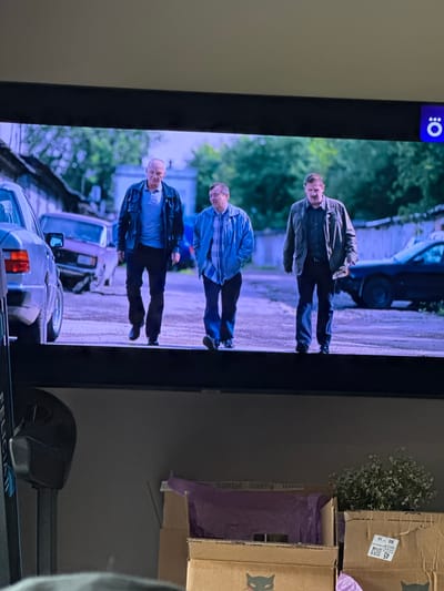 A photograph captures an indoor scene, primarily focused on a television screen positioned vertically. On the TV, a daytime outdoor scene is displayed, featuring three men standing on what appears to be a dirt road or path. The man at the top of the screen wears a dark jacket, a light shirt, and dark trousers, and appears to be smiling or talking. Below him, another man is dressed in a blue shirt and dark trousers. The third man, at the bottom of the screen, wears a dark jacket over a light shirt and dark trousers. Cars and lush green foliage are visible in the background of the screen. In the top left corner of the television, a white circular logo with a colon (:) inside, on a dark blue background, is visible; this is the Okko streaming service logo.
To the right of the television, two cardboard boxes are stacked. A small cluster of dried, light-colored flowers (resembling baby's breath) protrudes from the top box. A label on the top box shows numbers including "45," "80," and "10," along with a QR code. The word "FRESH" is faintly visible on the inner flap of the bottom box. At the very bottom of the image, part of a fitness machine, possibly a treadmill, is visible with the text "FELICIA II" partially legible. The immediate surroundings suggest an indoor setting, likely a home in Kuda, Russia, given the provided context. The room is dimly lit, contrasting with the bright, outdoor scene on the screen.