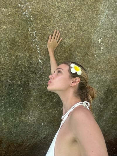 A fair-skinned woman lies on a rough, textured rock surface. A white and yellow flower is positioned in her hair above her left ear. She wears a white top with thin straps. Her left hand rests on the rock, displaying a ring on her ring finger. The location is likely near Lam Kaen, Thailand, given the tropical flora. The rock has a textured, mottled pattern with some white deposits along a linear path.
