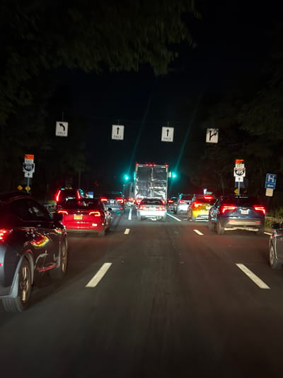 The image depicts a nighttime traffic scene on a highway, likely in or near the Village of North Hills, United States of America, given the presence of "EAST 495" signage. The highway has multiple lanes, and traffic appears to be moving slowly, suggesting a congestion. Several cars are visible, with their headlights and taillights illuminated. The road is paved and marked with white lane dividers. Overhead signs indicate lane usage: one shows an arrow pointing left with "ONLY" and another shows an arrow pointing straight up with "ONLY". Trees line the sides of the highway, and the sky is dark. A large semi-truck is also present in the traffic.