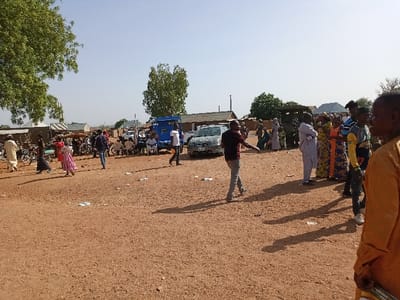The image depicts a scene in the city of Zum, Nigeria, located at coordinates 10.116 latitude and 13.046 longitude. The location is an open, dirt field with scattered debris. A large group of people, approximately 20 individuals, are gathered in the field. They are primarily standing, but some are seated on chairs, others are walking. Several vehicles are present, including a blue pickup truck and a silver car. The majority of individuals are wearing traditional clothing, indicating a possible community gathering or event. The people are interacting with each other, and some appear to be engaged in conversations. The environment is characterized by dry, dusty earth, suggesting a hot and arid climate. The sky is clear and bright, with little cloud cover. The scene appears to be taking place during the daytime. A large tree with green leaves is present on the left side of the frame, providing shade. There are also several smaller trees and bushes scattered around the area. The scene gives the impression of a public gathering or event, possibly in a village or town square. The relaxed demeanor of the individuals and the presence of vehicles suggest an environment of casual social interaction.
