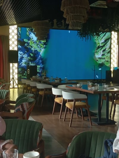 The image shows the interior of a restaurant or cafe, likely located in Бактуу-Долоноту, Kyrgyzstan. The dominant feature is a large, high-definition screen displaying a scenic view of a body of water surrounded by lush green terrain, possibly a lake or river with forested hills. The screen illuminates the space, casting a blue and green glow.
In the foreground, several tables are set for dining, with white tablecloths, plates, cups, and glasses. Chairs with white backs and mustard-yellow seats are arranged around the tables. To the left, there are plush green chairs with patterned accents, and further back, more dining tables and chairs are visible.
The ceiling features a grid-like structure and several large, tiered chandeliers made of a woven material, adding a rustic yet modern aesthetic. On the left side of the room, a tall speaker system is positioned next to a decorative panel with an intricate, illuminated geometric pattern. Curtains in a muted red tone hang on the far left. The flooring appears to be polished wood.
The overall ambiance suggests a contemporary dining establishment designed for comfort and visual engagement, with the large screen serving as a focal point. There are no visible people actively engaged in the scene, though the presence of dining settings implies recent or upcoming occupancy.