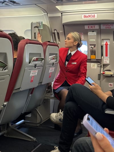 The image displays the interior of an aircraft cabin, located in the city of Novogorelovo, Russia.

A blonde female flight attendant, dressed in a red blazer, white collared shirt, and dark tie, is seated in the mid-ground, facing left and slightly upwards. Her right hand is extended towards an overhead panel or mechanism located above the emergency exit door. The exit door, positioned to her right, features both Cyrillic and English labels, including "ВЫХОД" and "EXIT," along with a red arrow.

In the foreground, the lower body of a passenger is visible, wearing dark trousers and white sneakers, with their legs crossed. This individual is holding a smartphone in their right hand. Adjacent to this, partially visible on the far right, is another passenger's hand holding a smartphone with a light purple case.

The cabin interior features rows of aircraft seats upholstered in red and grey, equipped with seatback pockets. The ceiling displays overhead compartments and lighting elements. The floor is covered with dark grey carpeting.