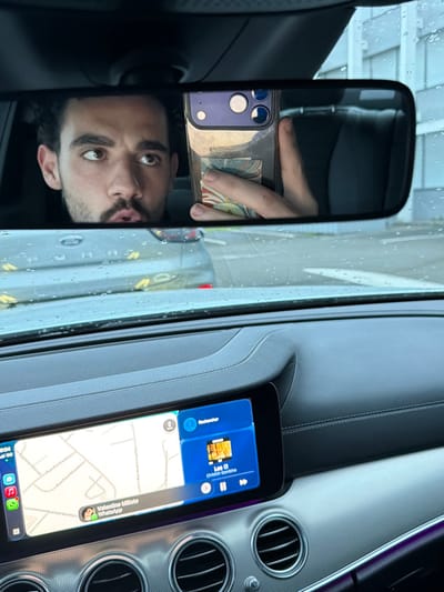 The image depicts the interior of a vehicle, viewed from the front passenger area, looking towards the windshield and rearview mirror. Water droplets are visible across the windshield, indicating rain or recent precipitation.

The rearview mirror reflects a male individual, likely the driver or front passenger, with dark hair, a beard, and brown eyes, looking towards the viewer's left with his mouth slightly agape. His right hand holds a smartphone, visible in the mirror, with a transparent case and a patterned back, indicating the individual is taking the photograph via the mirror's reflection.

Below the mirror, the car's dashboard features a wide digital infotainment screen. The left side of the screen displays a navigation map interface, showing street names including "Avenue de la Marne," "Rue Jean Baptiste Marcellin," "Rue Paul Maulong," and "Rue André Cabilis." These streets identify the location as Mont-de-Marsan, France. A WhatsApp notification from "Valentine Milleto" is visible at the bottom of the map display. The right side of the screen shows a media player interface, indicating the song "Les El" by "Chilouh Gambie" is currently playing, alongside a "Rechercher" (Search) option.

Below the screen, the dashboard features three circular air vents. The dashboard materials consist of dark grey and silver-grey textured surfaces, with a subtle purple ambient light visible on the far right. The scene outside the windshield shows the rear of a vehicle directly ahead.