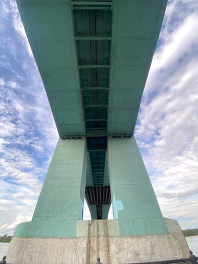 The media file presents an upward view from directly beneath a large bridge structure. The primary subjects are the bridge's robust undercarriage and the expansive sky. Massive, faded greenish-blue girders, likely constructed from metal or concrete, dominate the foreground, appearing to converge towards the center of the frame from this perspective. Beneath these main structural elements, a series of smaller beams and supports, some possibly wooden, form a more intricate network.

On the far right of the image, a substantial light-gray concrete abutment or pier supports the bridge. A railing or fence-like structure is faintly visible along the top edge of this abutment. The background is filled with a bright blue sky dotted with scattered white, cumulus clouds, indicating it is daytime with partially cloudy weather. In the very far background, beyond the concrete pier, a narrow strip of green land (suggesting trees or foliage) and a glimpse of water are discernible, implying the bridge spans over a natural landscape feature.

No human activity is depicted. On the concrete abutment on the right, faint, white vertical markings or symbols are present, but they are too indistinct and partially obscured to be clearly read or identified. The location context for this scene is Grigor'evskoe, Russia.