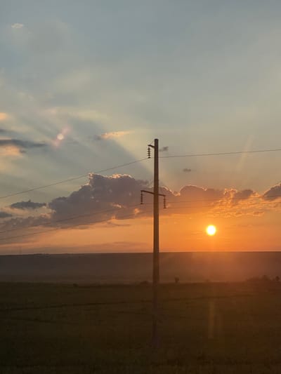 The image depicts a sunset scene in Balabanu, Moldova. A single utility pole stands in the foreground, supporting electrical wires that stretch across the frame. The sun, a bright orange orb, is setting on the horizon, casting a warm glow across the sky and illuminating scattered clouds. A vast, dark field stretches out below the horizon, suggesting agricultural land. The overall impression is one of rural tranquility at dusk.