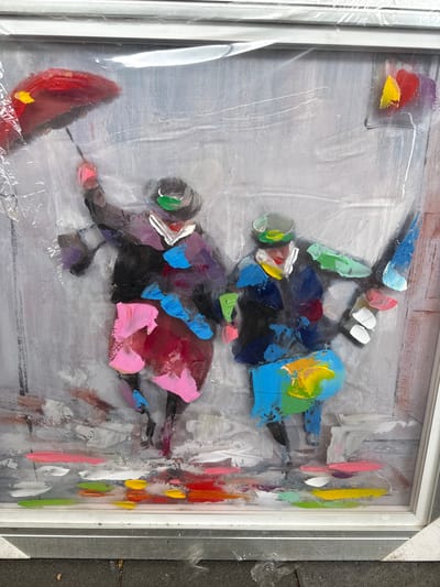 The image shows a framed abstract painting with thick brushstrokes. The painting depicts two figures on horses, with the upper figure in blue and yellow and the lower figure in pink and black. The background is a mix of gray and white. The frame is white. The painting appears to be in Aubange, Belgium. The image is slightly obscured by a layer of plastic wrap on the frame.