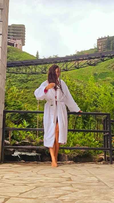 A woman stands on a flagstone terrace, wearing a white robe and facing slightly to the right. She is barefoot and rests her left hand on the black metal railing in front of her. The backdrop includes a lush green hillside and a bridge with an abandoned building on the crest of the hill. A small waterfall spills over rocks just beyond the railing. The location is Cat Cat Village, Vietnam.