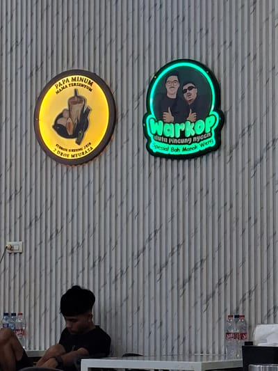 The image shows two illuminated signs on a vertical striped wall. The sign on the left is circular with a yellow glow and depicts a beverage and some food items. Text above reads "PAPA MINUM MAMA TERSEYUM" and below it, "STROKE 2 DROE MEURASA". The sign on the right is a rounded rectangle with a green glow and features a cartoon illustration of two men. Text within the green glow reads "warkop" and "duta pincung nyeeen spesial Bah Manok Weng".
In the foreground, a young man with dark hair and a black t-shirt is sitting with his arms crossed, looking down. He is seated at a white table. To the left of the man, there are three bottles of water on the floor. To the right of the table, there are two more bottles of water and a white tissue. The wall has a textured, ribbed pattern with dark grey and white marbling. The environment suggests an indoor setting, possibly a cafe or restaurant. The text on the signs indicates that the establishment is located in Sigli, Indonesia, a city in the Aceh province.