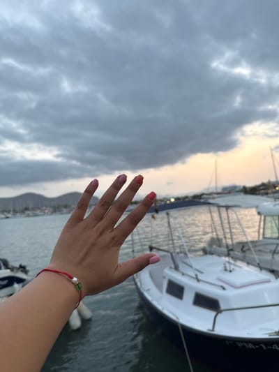 A person's hand with painted nails is held up in front of a marina scene. The hand, with pink and orange nail polish, has a red string bracelet with a pearl and green bead on the wrist. In the background, several boats are docked in the water. The sky is cloudy with a hint of sunset glow visible on the horizon. The location is Port D'alcúdia, Spain.