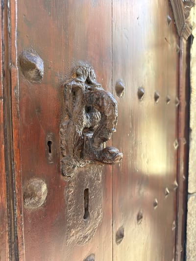 This is a close-up, eye-level, outdoor, full shot of an ornate, weathered, dark wooden door in Tossa de Mar, Spain. The door has a rough, aged texture, with visible wood grain and a glossy finish that reflects the ambient light. It is studded with numerous small, diamond-shaped metal rivets, giving it a rustic and fortified appearance. To the left of the center, a prominent, intricate, wrought-iron door knocker is attached. The knocker is shaped like a stylized animal, possibly a dragon or lion, with detailed facial features and a coiled body. Below the knocker, a dark, keyhole-shaped lock plate is visible. The stone wall of a building is visible to the far right of the frame, framing the door. The overall impression is one of historical significance and robust craftsmanship.