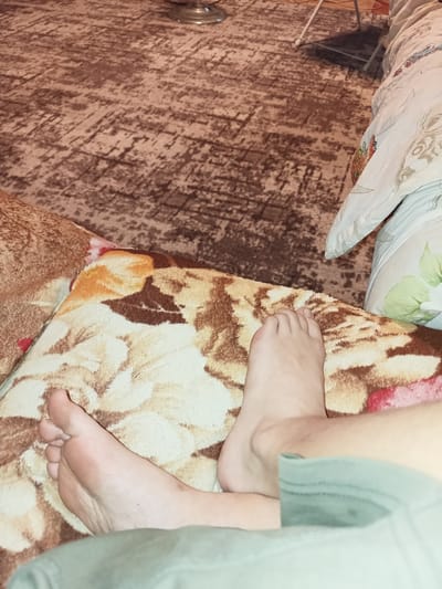 The image shows a close-up view of two feet resting on a patterned blanket. The blanket has a floral design with shades of brown, cream, and red. In the background, a carpet with a similar color palette and an abstract pattern is visible. Part of a pillow with a subtle design can also be seen. The lighting suggests it might be indoors, possibly in a living room or bedroom. The overall scene appears relaxed and casual, with the feet positioned comfortably. No specific activity is clearly depicted, other than resting. There is no visible text in the image. The location context of Temernitskiy, Russia, does not provide any immediate visual cues within the image itself.