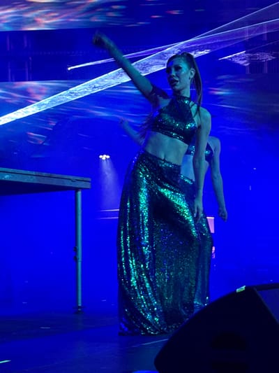 In Akhalubani, Georgia, on a dimly lit stage, two female dancers perform under intense blue lighting and laser effects. The dancer in the foreground, facing the audience, wears a shimmering green sequined halter top and a matching floor-length skirt, her hair pulled back in a ponytail. She is captured mid-performance, with her left arm extended upwards. Behind her, another dancer, also in a similar green sequined outfit, is partially visible, her face obscured. The stage floor is dark, and a silver platform with cylindrical legs is positioned to the left of the main dancer. The background is a vibrant blue, illuminated by bright laser beams that cut across the scene, creating a dynamic and energetic atmosphere.