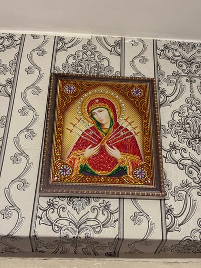 A framed diamond painting of a religious icon hangs on a wall covered in patterned wallpaper. The diamond painting depicts the Virgin Mary, adorned in a red robe and blue headscarf, with a halo around her head. Seven swords pierce her chest, and her hands are clasped in front of her. The wallpaper has a light background with a repeating dark grey floral and scroll pattern. The painting is mounted centrally on the wall, above a white baseboard. The lighting appears to be artificial, casting a shadow on the baseboard below. The scene is indoors.