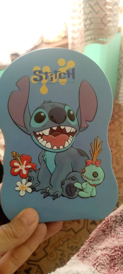 The image displays a light blue, heart-shaped object featuring the cartoon character Stitch. Stitch is depicted with his mouth wide open, a red tongue visible, and numerous sharp white teeth. He has large, round blue eyes and prominent purple ears. To the left of Stitch are two hibiscus flowers, one red and white, the other white and yellow. To Stitch's right is a small, green voodoo doll character with black button eyes, a red bow, and yellow grass-like hair. The object is held by a hand, with the thumb and index finger visible at the bottom left. The background is blurred and consists of textured fabrics and a mint green wall. There is no indication of the city of Marhamat, Uzbekistan in the image.