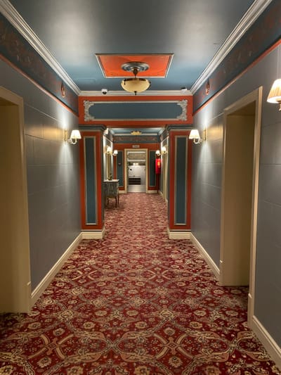 A long hallway with intricate design elements. The left wall is blue with framed artwork and sconces. The right side is covered in a dense red patterned carpet. Overhead, the ceiling is segmented by beige beams. Arched doorways lead deeper into the hall, illuminated by ornate wall lamps. The hallway floor is tiled in a muted gray.
