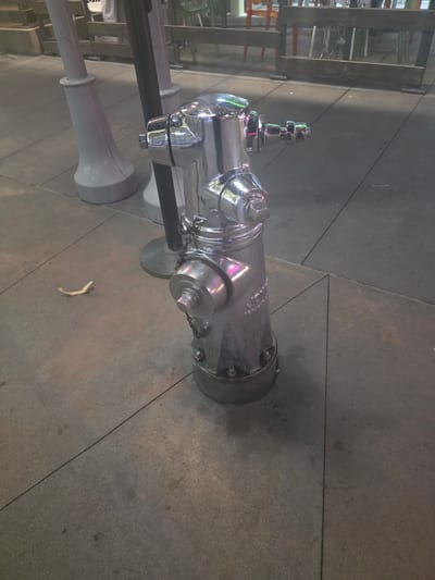 A gleaming chrome fire hydrant stands on a paved sidewalk in Burbank, California. The hydrant is a bright silver, reflecting the ambient light. It has a traditional design with several caps and nozzles. The sidewalk is made of large, dark gray rectangular concrete slabs, with visible seams between them. In the background, a thin black pole is visible, and beyond that, a wooden fence with chairs and tables suggests an outdoor seating area, possibly for a restaurant or cafe. There are also some light-colored, rounded posts or bollards on the left side of the image. A small, light-colored piece of paper is on the ground near the base of the hydrant. The overall scene appears to be an urban outdoor environment.