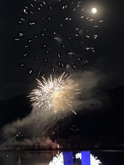 This media file depicts a nighttime fireworks display. The scene is dark, indicating it is likely late evening or night. The main subjects are the bright, explosive bursts of fireworks against the dark sky. There are multiple fireworks visible, with some appearing as starbursts of light and others as trails of sparks. Smoke from the explosions drifts through the air, obscuring some parts of the scene.
On the left side of the image, there is a large, bright, circular object which could be the moon or a very bright light source. Scattered around this object are numerous smaller, bright specks of light, which are likely more fireworks or their remnants.
In the center and right side of the image, a large, vibrant fireworks burst is prominently displayed. This firework is characterized by its many radiating lines of light and a central bright core, surrounded by smoke.
To the far right, there are some indistinct structures that appear to be illuminated. Two rectangular areas of bright blue light are visible, possibly part of a building or display. Above and below these blue rectangles, there are white bursts of light, suggesting more fireworks or festive lighting. A dark, vertical structure runs alongside these illuminated elements, possibly a building or a pole.
The overall impression is one of a celebratory event, likely a festival or holiday, given the presence of fireworks. The setting appears to be outdoors, possibly near water or an open area to accommodate such a display. There is no visible text in the image. The time of day is clearly night. The weather is not explicitly indicated, but the clear visibility of the fireworks suggests it is not raining or heavily overcast.