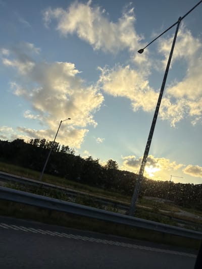 The image depicts a sky with scattered clouds, illuminated by a setting or rising sun. Sunbeams are visible radiating from behind the clouds. In the foreground, a dark road with white lane markings is visible, separated from a grassy verge by a metal guardrail. A tall street lamp with a curved arm and a dark fixture stands prominently on the right side of the frame, silhouetted against the sky. Another street lamp is visible further in the background on the left. A dense line of dark trees forms a backdrop behind the grassy verge. The overall impression is of a roadside scene during twilight. The location is Ödsmål, Sweden.