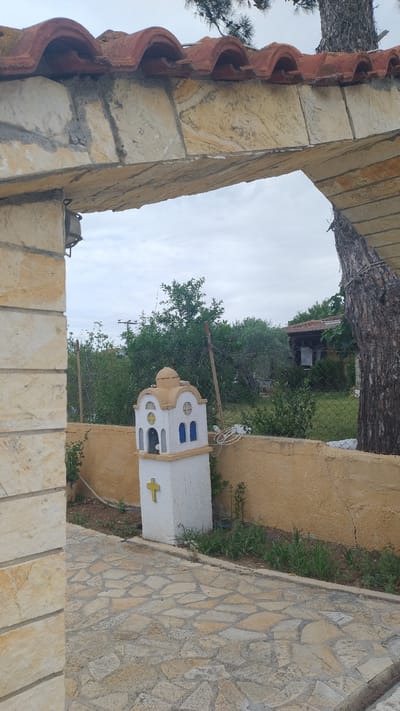 The image shows an outdoor scene viewed through a stone archway with a tiled roof. In the foreground, a pathway made of irregularly shaped stones leads towards the interior of a courtyard. To the left, a stone pillar supports the archway. In the middle ground, a small, white, steeple-like structure with a dome is visible, featuring blue arched windows and a yellow cross on its side. This structure is situated in front of a yellow stucco wall. Behind the wall, a fence separates the courtyard from a garden area with lush green trees and vegetation. A rustic building with a tiled roof is partially visible in the background. The sky is overcast, and a large tree trunk is prominent on the right side of the image, leaning into the archway. The scene appears to be in Νέα Κερασιά, Greece, likely a residential or religious site given the presence of the miniature church-like structure. There are no people or animals visible in the image.