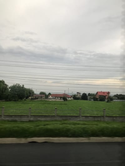 This detailed image captures an outdoor scene in Răcăciuni, Romania, presenting a semi-rural or suburban landscape under an overcast sky. The upper two-thirds of the frame are dominated by a cloudy, light grey sky with darker cloud formations present in the lower sky, particularly towards the right where a brighter patch hints at light filtering through. Multiple parallel utility lines, approximately seven, traverse horizontally across the mid-upper section of the frame.

The foreground, approximately the bottom 15% of the image, shows a dark, asphalt-like paved surface, likely a road or pathway, exhibiting a slight motion blur. Above this, a light-colored concrete curb or low barrier extends horizontally, followed by a narrow strip of dark green grass.

The midground features a large expanse of vibrant green grassy field, appearing lush and well-maintained. A low fence runs horizontally across this field, positioned roughly one-third of the way up from the bottom of the image. This fence is composed of light-colored vertical posts, with at least five clearly visible, connected by one or two horizontal rails.

The background reveals scattered structures and dense vegetation. On the far right, a prominent house with a distinct reddish-orange tiled roof and light-colored walls is partially visible. Towards the center-left, another building with a similar reddish roof and light walls is partially obscured by trees. Farther to the left, additional smaller structures are discernible. In the distant center-right, a taller, blocky, possibly industrial structure or water tower is faintly visible. Numerous dark green deciduous trees, varying in size and density, are interspersed throughout the background, providing cover for some buildings and forming natural clusters along the horizon line. No people, animals, or active interactions are visible in the scene.