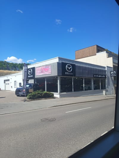 The image shows a Mazda dealership in Nancy, France, on a bright, sunny day. The dealership is a modern building with large glass windows displaying several Mazda cars. A black Mazda SUV is parked in front of the dealership, with a large Mazda flag to its left. The street in front of the dealership is paved and has a white lane marking. In the background, there are trees and a blue sky with some clouds. The dealership's name, "Pasion Automobile", is visible above the windows.