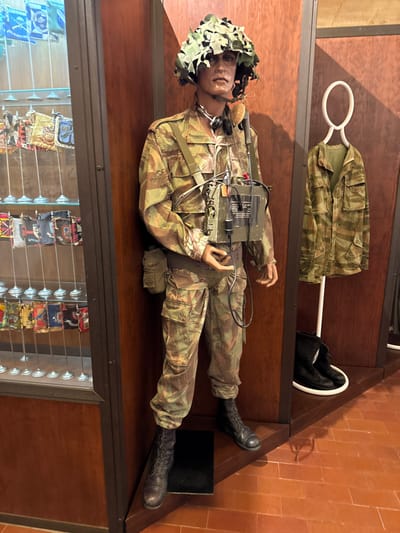 The image depicts a museum exhibit located in Praia do Ribatejo, Portugal. The central subject is a full-size mannequin dressed in a complete military camouflage uniform featuring a light green, brown, and dark green "lizard" or "tiger stripe" pattern. The mannequin wears a dark green helmet covered with a camouflage net, a headset with an earpiece and microphone, and has a rectangular, dark green military radio or communication device strapped to its chest with visible cables and red markings. A dark green belt with a pouch is secured at the waist, and black military boots complete the attire.
To the mannequin's immediate right, another camouflage jacket of the same pattern hangs from a white hanger on a white vertical stand. Below it, on the terracotta-tiled floor, a pair of black military boots rests on a white circular display base.
To the mannequin's left, a tall, glass display cabinet with dark wooden framing showcases multiple rows of small, decorative flags or pennants, each mounted on a thin metal pole with a circular base. The flags exhibit various colors and emblems. The overall environment features dark wooden paneling and a terracotta-colored tiled floor.