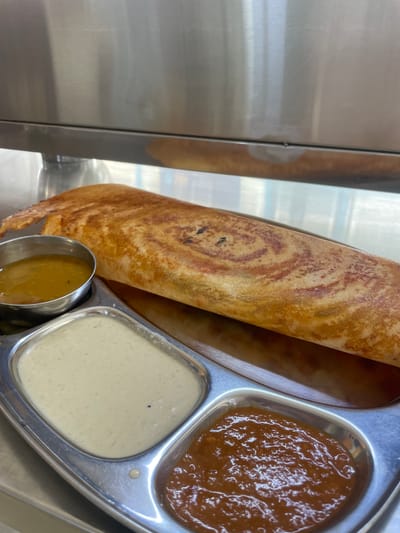 A close-up, eye-level, overhead shot displays a South Indian meal served on a metal thali tray. The meal consists of a rolled dosa, a thin savory pancake, golden brown with reddish-brown swirling patterns, laid diagonally across the tray. To the left of the dosa, there is a small steel bowl containing a yellow, liquid sambar. Below this, in a larger compartment of the thali, is a creamy white chutney. To the right of the white chutney, in another compartment, is a reddish-brown chutney. The background is a plain, reflective stainless steel surface, likely a countertop or serving area. The lighting is even and bright, highlighting the textures and colors of the food. The scene appears to be from a restaurant in Kurnool, India, given the typical presentation of South Indian cuisine.