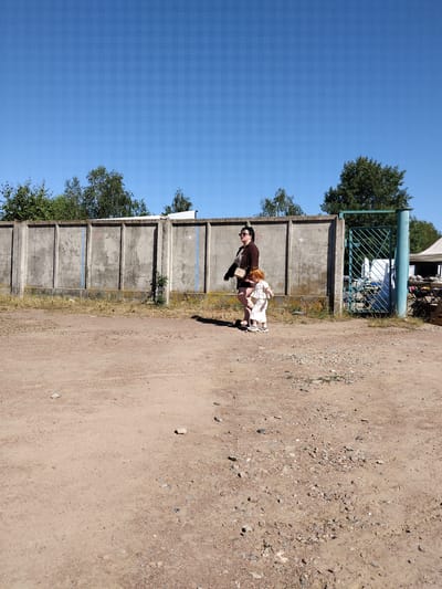 A woman and a young child stand on a dirt path in front of a tall, grey concrete wall with vertical divisions. The woman, on the left, wears a dark brown jacket over a striped shirt and dark shorts, with sunglasses and a light-colored crossbody bag. The child, to her right, has red hair and wears a white outfit. To the right of the wall, a green metal gate is visible. Beyond the gate, a table with items for sale is set up under a white canopy. Trees line the top of the wall and extend beyond it. The ground is covered in dirt and small stones, with sparse, dry grass growing along the base of the wall. The sky is clear and blue. The scene appears to be an outdoor market or storage area in Irshansk, Ukraine.