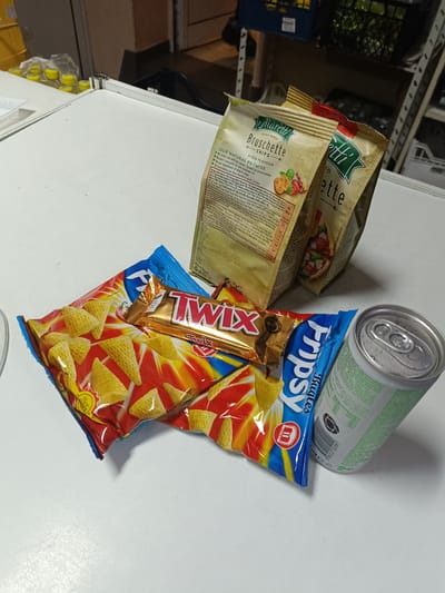 The image shows a flat, white surface upon which various food items are placed. There are two bags of "Marelli Bruschette Chips" of pizza flavor, one "Twix" chocolate bar, a bag of "Fripsy Bugles" chips, and a can of soda. The packaging labels indicate the chips are made in Bulgaria. The location "Pripek" is not visible in the image.
