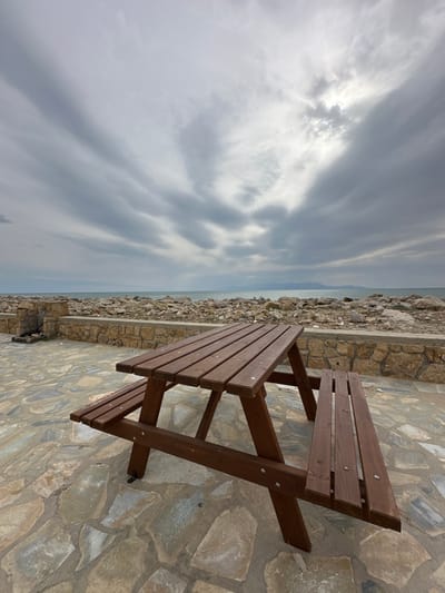An outdoor scene in Limenas Avdiron, Greece, features a brown wooden picnic table with attached benches positioned on a flagstone-paved area in the foreground. The table is oriented with its length parallel to the viewer, slightly angled towards the right. Beyond the paved area, a low stone wall constructed from irregular light-colored stones separates the foreground from a rocky shoreline composed of numerous large, light-colored boulders. The sea extends horizontally across the mid-ground, appearing calm under an overcast sky. Distant landmasses, possibly mountains, are visible on the horizon line across the water. The sky, occupying the upper two-thirds of the image, is heavily clouded with various shades of grey, and brighter patches of light are visible where sunlight partially penetrates the cloud cover. No individuals or active subjects are present within the frame.