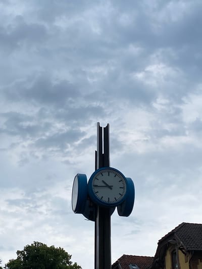 The image shows a clock with blue casing mounted on a dark metal bar, against a cloudy sky. The clock face is white with black markings and hands. The hour hand points to approximately 9, and the minute hand points to approximately 45. Below the clock, a second, identical clock casing is visible, also mounted on the metal bar. To the right, the top of a building with a dark tiled roof and yellow facade is visible, suggesting an urban or town setting. In the lower right corner, the top of a lush green tree is visible. The sky is overcast with numerous grey clouds, indicating a cloudy or possibly stormy day. The image is taken from a low angle, looking upwards. The location is identified as Rinteln, Germany. No people are visible, and no text is discernible on the clock or in the scene. The overall impression is of an overcast day in a town, with a prominent public clock.