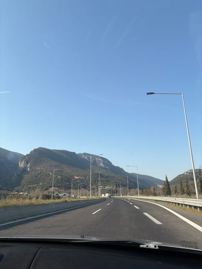 The image depicts a forensic view of a multi-lane highway cutting through a mountainous, vegetated landscape under a clear blue sky, as seen from the interior of a moving vehicle. The location is the Δημοτική Ενότητα Ευρυμενών, Greece.

The foreground shows the dark-colored dashboard and part of the windshield of the observer's vehicle. The asphalt highway extends into the distance, featuring at least two lanes of travel marked with clear white dashed and solid lines. To the immediate right, a paved shoulder is visible, bordered by a concrete barrier topped with a metal guardrail. On the left, a concrete median barrier separates the opposing lanes.

Numerous tall, slender streetlights with horizontal arms and rectangular luminaires line both sides of the highway, receding into the background. The environment is dominated by large, steep mountains covered in dense, dark green vegetation on both sides of the road. In the mid-ground on the left, several tunnel-like structures are visible at the base of the mountains, along with what appear to be overhead gantries carrying traffic signals or signs. A few distant vehicles are discernible on the highway ahead, indicating active traffic. The sky is uniformly light blue with minimal cloud cover. No individuals are explicitly visible.