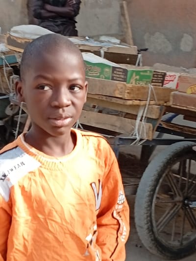 This image depicts an outdoor scene in Gayawa, Nigeria. A young male subject, approximately 8-12 years of age, with dark skin and a close-cropped haircut, occupies the foreground, looking slightly towards the viewer's right with a neutral expression. He wears an orange long-sleeved shirt featuring white and grey accents on the shoulders and sleeves, including a basketball motif on the right sleeve. In the upper left background, a portion of an adult male in dark attire is visible, appearing to be seated or crouched with arms crossed. The midground contains a structure composed of stacked wooden pallets or planks, upon which various cardboard boxes are placed. Identifiable text on one box reads 'M-PLUS' on a green background. To the far right, a spoked wheel with a dark tire is visible, consistent with a cart or bicycle. The immediate environment includes an uneven ground surface and a light-colored, weathered wall in the distant background. The scene is under natural daylight conditions.