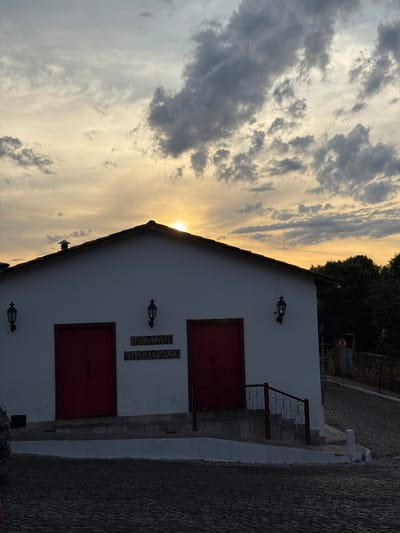 The image captures the "Temperod Rosário" restaurant in Pirenópolis, Brazil. The white building, with two sets of red doors and matching wall-mounted lamps, is situated against a backdrop of a sunset-filled sky with dramatic clouds. A sign above one of the doors identifies the building as a "Restaurante Temperod Rosário." Steps lead up to the doors, with a simple metal railing on one side. A section of cobblestone pavement is visible on the right side of the image. In the top right corner, a no parking sign on a wooden post is partially visible.