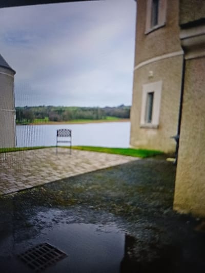 The image depicts an outdoor scene at the Lough Erne Resort in Enniskillen, County Fermanagh, United Kingdom, overlooking Lough Erne under an overcast sky. In the foreground, a dark, wet gravel or loose stone surface with a prominent puddle occupies the right and lower left. A square metal grate is visible within the puddle in the bottom left corner. To the left of the wet area, a paved patio section with light-colored, rectangular pavers laid in a herringbone or similar pattern is present. A single dark-colored, possibly metal, outdoor chair with an open-patterned backrest stands centrally on this paved area, facing the water.
To the left, part of a light-colored, rounded building or tower structure is visible. To the right, a substantial building with light-colored rendered walls features a rounded section with two narrow, vertical, recessed windows with light frames. A dark vertical drainpipe runs down the right side of this building. Between the paved area and the building on the right, a strip of green grass is visible.
In the mid-ground, a wide expanse of calm water, identified as Lough Erne, stretches horizontally. Beyond the lough, a distant treeline and undulating green landscape are visible under a uniformly cloudy, bright grey sky. No individuals or active subjects are present. The scene conveys a quiet, damp, and unoccupied waterside environment.