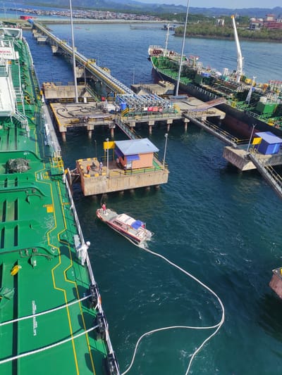 This elevated shot captures a bustling port scene in Kunak, Malaysia, under clear, bright daylight. The main subject is a large, green-hulled tanker, likely an oil or chemical carrier, either moored or in the process of mooring at a substantial concrete pier.

The tanker's green hull is marked with yellow lines and displays text such as "F4," "WB7S," and "COSSORSCOORS SLOPS." A "NO SMOKING SAFETY FIRST" sign is also visible on its side. The tanker's deck is a network of pipelines, hatches, and a white crane arm. Several thick white mooring lines extend from the tanker's side, securing it to the pier, with one line running into the water from a smaller vessel.

Alongside the tanker, a long concrete pier extends into the blue-green water. This pier is connected to the distant land by an extensive jetty, which is lined with numerous pipelines, indicating an industrial facility. On the pier itself, there's a small structure with a blue roof and various concrete platforms.

A smaller white and red pilot boat, clearly marked with "PILOT" on its side, is positioned close to the tanker. This boat appears to be assisting in the mooring operations, with a heavy white rope trailing from its stern into the water towards the tanker.

Several people are visible in the scene: individuals in work attire are on the tanker's deck, handling mooring lines, and others are on the pier and the pilot boat, indicating active operations.

In the background, the coastline features green, rolling hills and a densely packed settlement of buildings stretching along the water, suggesting a populated coastal area. The water is relatively calm, reflecting the clear sky.