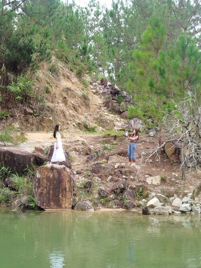 In Lán Tranh, Vietnam, two young women are positioned in an outdoor natural environment. The woman on the left, with long black hair, stands on a large rock, wearing a yellow top and a white skirt. The woman on the right, with long brown hair, stands on the rocky terrain, holding a phone and seemingly taking a photo. The landscape is characterized by a body of green water in the foreground, with a steep, rocky embankment rising behind it. Dense green pine trees cover the upper portion of the embankment and the background. The ground is covered with loose rocks and dirt, with some sparse vegetation.