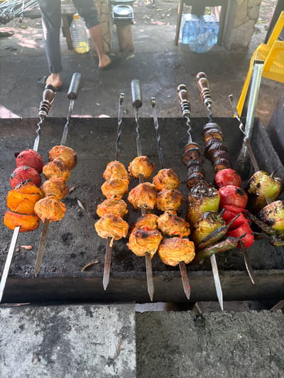 The image depicts an outdoor cooking scene, likely in Abzhanda, Abkhazia, with a focus on grilled food. In the foreground, several skewers of food are arranged on a dark, presumably charcoal, grill. These skewers contain a variety of items, including what appear to be meatballs or kofta, grilled tomatoes, bell peppers (green, red, and yellow), and possibly pieces of eggplant. The food shows signs of being cooked over heat, with some charring and browning.
In the background, out of focus, are elements suggesting a casual, outdoor environment. There are glimpses of people's legs and feet, wearing sandals and casual clothing. A large plastic bottle filled with a light-colored liquid, possibly cooking oil or water, is visible. Other elements of the environment include what appears to be wooden structures or furniture, and a bright yellow object, possibly a chair. The ground surface seems to be a mix of dirt and concrete. The overall impression is a relaxed, informal setting where food is being prepared outdoors.