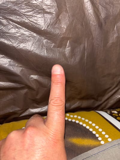 A close-up, eye-level shot shows a person's index finger, with a slight reddening on the tip, pointing upwards against a crumpled, dark brown, textured background. The textured background appears to be a plastic tarp or similar material, with a pattern of small raised dots. In the lower portion of the image, a yellow and brown patterned fabric is visible, with a gray fabric band at the bottom edge. There are no identifiable landmarks or context that would suggest a specific location within the city of Gribki, Russia. The image is well-lit, with the primary focus on the finger and the texture of the material behind it.