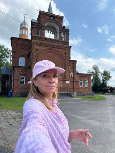 A blonde woman with freckles, wearing a pink and white striped shirt and a pink baseball cap with an embroidered logo, stands in front of a red brick church in Chudovo, Russia. She is looking towards the camera, with her right hand resting on her chest. The church has a bell tower with arched openings and a silver dome topped with a cross. The sky is cloudy with patches of blue. There is green grass and a paved area in front of the church.