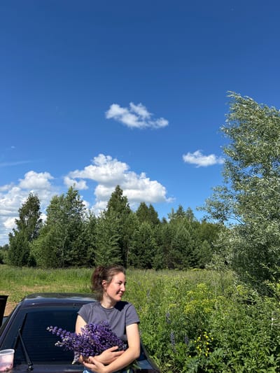A woman with dark hair tied back in a ponytail, wearing a grey t-shirt, stands next to a dark-colored car with her left side towards the camera. She is holding a large bouquet of purple lupine flowers in her arms and looking to her right. The car's rear window and windshield wiper are visible. To the right of the car is a field of green grass with various wildflowers, including yellow and purple ones, and taller green plants. Behind the field, a dense line of various green trees forms a backdrop, with a very tall, light-colored leafy tree on the far right. The sky above is a clear, bright blue with scattered white cumulus clouds. The scene is outdoors on a sunny day, likely in the village of Verkh-Rozhdestvo, Russia.