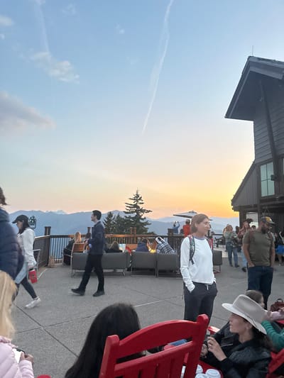 The image captures an outdoor patio scene at sunset, likely situated at a lodge or facility on Aspen Mountain, Colorado, providing a panoramic view of distant mountain ranges. Approximately fifteen to twenty individuals are visible. In the foreground, a red wooden rocking chair partially obscures a person with dark hair and a woman in a light-colored hat and dark jacket. Centrally, a woman in a white long-sleeve top and dark pants stands with hands in pockets, looking left, while a man in a dark suit walks from left to right. To their right, a man in a green t-shirt and baseball cap stands. In the midground-background, multiple groups are seated on grey sofas surrounding a circular fire pit with visible flames, socializing. Other individuals are standing or walking across the light-colored paved patio. A dark railing separates the patio from a forested slope leading to a valley and blue-hued mountains on the horizon. A tall evergreen tree stands prominently behind the fire pit. To the right, a multi-story, dark-colored lodge-style building with a steep roof is present. The sky above transitions from orange and yellow at the western horizon, indicative of sunset, to pale blue, with a distinct contrail diagonally traversing the upper left.