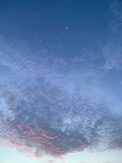 The image captures a sky scene with a crescent moon visible in the upper portion. The sky is a deep blue, transitioning to lighter hues towards the bottom. Scattered across the sky are small, white, puffy clouds, with some areas showing a soft pinkish hue, suggesting the presence of twilight or sunrise/sunset lighting. A larger mass of these textured clouds occupies the lower half of the frame, with the pinkish coloration more pronounced in this area. The overall impression is of a serene sky. This scene could be observed from anywhere in Karandugoda, Sri Lanka, given it is an outdoor sky view. No people, subjects, objects, actions, or interactions are depicted. The environment is the sky itself.