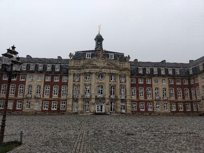 The image depicts the Schloss Münster (Münster Palace) in Münster, Germany, under an overcast sky. The large, symmetrical Baroque-style building, positioned centrally, presents a facade of red brick and light-colored stone. A prominent, elaborately decorated central section features multiple arched and rectangular windows, decorative columns, sculptural elements, and two main entrance doors at ground level. This central section is topped by a green-domed cupola with a golden spire and an ornate golden finial. Symmetrical wings extend from the central structure, exhibiting a pattern of red brick walls contrasted with light stone window surrounds and decorative elements. The building has a dark grey mansard roof dotted with numerous dormer windows.
In the foreground, a vast, empty courtyard is paved with dark grey cobblestones, forming distinct linear patterns leading towards the building. A dark, ornate, multi-lantern streetlamp is partially visible on the far left, entering the frame from the bottom. A small, grey metal bollard is situated in the lower left foreground, next to a patch of green grass. No individuals, vehicles, or specific activities are present within the scene. The overall lighting is diffuse due to the uniformly grey, cloudy sky.