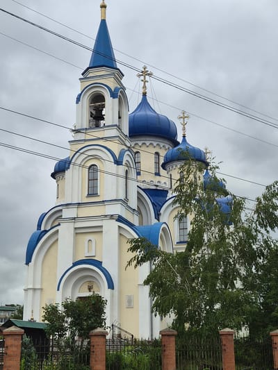 An Orthodox church with blue domes and a golden spire is visible under a cloudy sky. The church is painted yellow and white with blue trim. There are three bells hanging in the belfry. In front of the church, there is a metal fence and some trees. Power lines are visible crisscrossing the sky in front of the church. The church is located in the city of Arseniev, Russia.