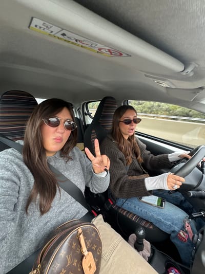 Two females are depicted inside a vehicle in motion, located in Le Cannet-des-Maures, France. The passenger, seated on the left, has long dark hair, wears reflective aviator-style sunglasses, a grey long-sleeved sweater, and a fastened seatbelt. She is shown with a puckered mouth expression and displays a V-sign (peace sign) with her right hand. A brown Louis Vuitton monogram canvas bag rests on her lap, partially obscuring beige trousers. The driver, positioned on the right, also has long dark hair, wears dark oval sunglasses, a dark brown knit sweater over white long sleeves, and blue jeans, with a fastened seatbelt. Her hands are on the steering wheel, and her mouth is open, suggesting speech or singing. A mobile phone lies on her lap. The car's interior features vertically striped seat upholstery and a manual gear shift lever between the front seats. The external environment, visible through the windshield, is blurred, showing a road alongside a green, tree-lined embankment. An airbag warning sticker is affixed to the headliner above the passenger side.